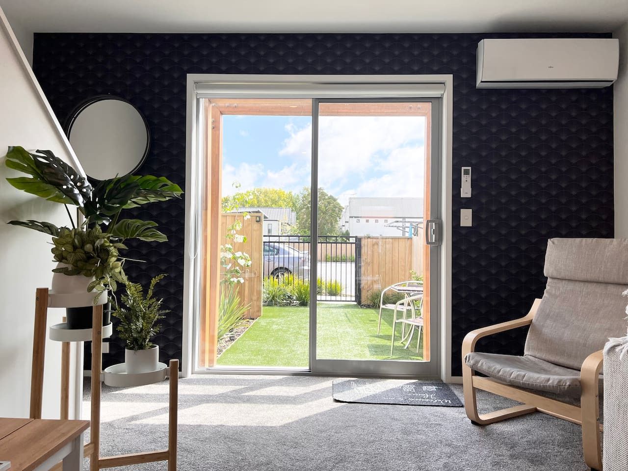 Ein modernes Wohnzimmer mit einer Schiebetür zu einem Garten. Es gibt ein graues Sofa, einen Holzstuhl und einen Blumentopf.