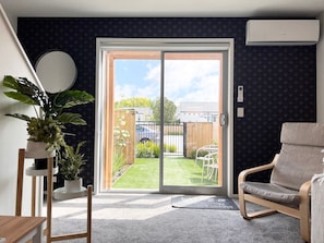 A modern living room with a sliding glass door leading to a backyard. There is a grey sofa, a wooden chair, and a potted plant.