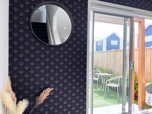 Un interior moderno con una pared oscura de diseño, un espejo redondo y un puerta corredera de cristal que da a un patio trasero con una cerca de madera y muebles de exterior.