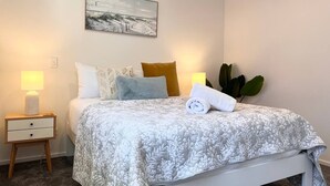 Een net opgemaakt bed met een beddekleed met patroon, een kussen, een opgerolde handdoek en een bedlampje op een houten nachtkastje.