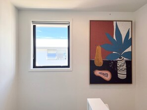 Um quarto com janela, um quadro e uma prateleira branca.