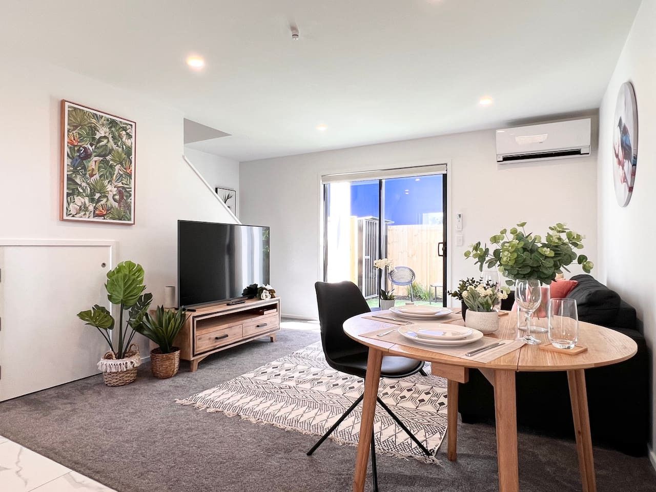 A modern living room with a dining table, a flat-screen TV, and a staircase leading to a balcony.