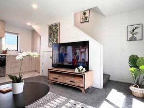 A modern living room with a flat-screen TV on a wooden stand, a dining table with a white orchid, and a staircase with a plant on the landing. - Salisbury townhouse with courtyard and workspace in Christchurch (Christchurch)