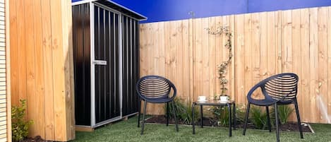 Un pequeño espacio exterior con césped artificial, dos sillas negras, una mesita con una taza y platillo, y un cobertizo de madera.
