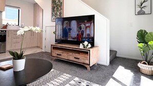 Ein modernes Wohnzimmer mit einem Flachbildfernseher auf einem Holzboden, einem Esstisch mit einer weißen Orchidee und einer Treppe mit einer Pflanze auf der Zwischenebene.