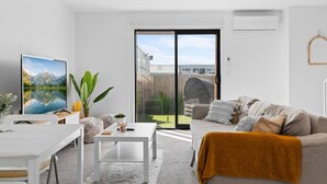 Un salon moderne doté d’un téléviseur à écran plat, d’un canapé, d’une table basse et offrant une vue sur un jardin avec une terrasse en bois et un espace salon extérieur abrité.