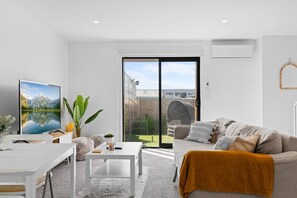 A modern living room with a flat-screen TV, a sofa, a coffee table, and a view of a backyard with a wooden deck and a covered outdoor seating area. - Sunny townhouse with parking and EV charging in Christchurch (Christchurch)