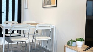 Une pièce moderne avec une table et des chaises blanches, une œuvre d’art encadrée au mur et une petite plante posée sur une table d’appoint.