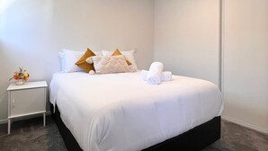 Un lit bien fait, avec du linge de lit blanc et quelques coussins décoratifs, une petite table de chevet sur laquelle est posé un vase de fleurs, et une porte fermée en arrière-plan.