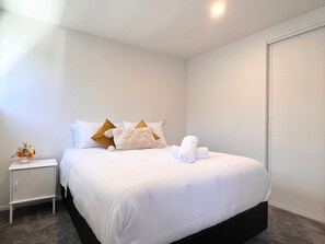 A neatly made bed with white bedding and a few decorative pillows, a small bedside table with a vase of flowers, and a closed door in the background. - Sunny townhouse with parking and EV charging in Christchurch (Christchurch)