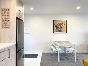 Une cuisine moderne avec un réfrigérateur en acier inoxydable, des meubles blancs et une table à manger avec des chaises. Un tableau encadré est accroché au mur, au-dessus de la table.