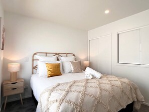 A neatly made bed with white and gold bedding, a bedside lamp, and a small wooden nightstand with drawers.