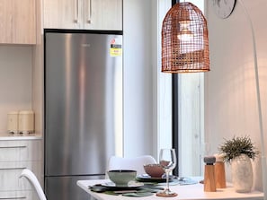 A modern kitchen with a stainless steel refrigerator, a white dining table with a glass top, and a hanging orange wire light fixture.