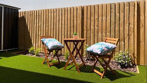 Eine Terrasse mit zwei Holzstühlen und einem kleinen Tisch, umgeben von einem Holzzaun und einer Rasenfläche.