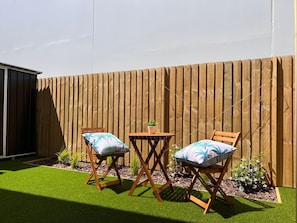 A patio with two wooden chairs and a small table, surrounded by a wooden fence and a grassy area.