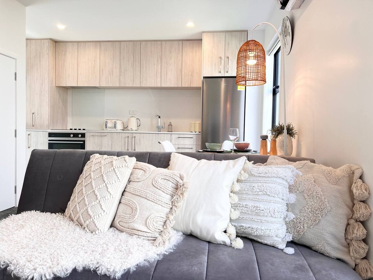 A modern living room with a grey sofa, multiple throw pillows, a fluffy white rug, and a kitchen in the background.