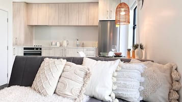 A modern living room with a grey sofa, multiple throw pillows, a fluffy white rug, and a kitchen in the background.