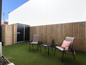 A patio with artificial grass, two chairs, a small table, and a wooden fence.