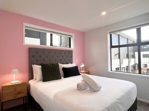 A hotel room with a large bed, a headboard, a nightstand, a lamp, and a window with a view of buildings.