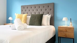 Un lit bien fait, doté d’une tête de lit capitonnée grise, de draps blancs et de plusieurs oreillers de différentes couleurs. On y trouve une table de chevet en bois, sur laquelle sont posés une lampe et une bouteille.