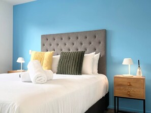 Una cama bien hecha con un cabecero gris con capitoné, ropa de cama blanca y múltiples almohadas de varios colores. Hay una mesita de noche de madera con una lámpara y una botella encima.
