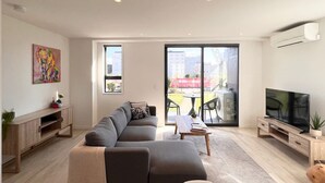 Een moderne woonkamer met een grijze modulaire bank, een salontafel, een televisie op een houten dressoir en een schuifpui die leidt naar een buitenruimte met een tafel en stoelen.