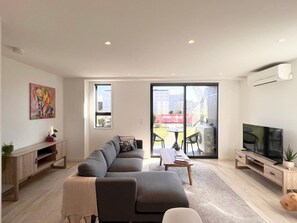 A modern living room with a grey sectional sofa, a coffee table, a TV on a wooden stand, and a sliding glass door leading to an outdoor area with a table and chairs.