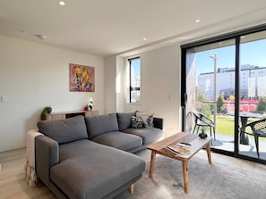 A modern living room with a grey sectional sofa, a wooden coffee table, and a view of the outdoors through large windows.
