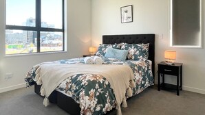 Een slaapkamer met een groot raam, een bed met bloemenbeddelsels, een nachtkastje met een lamp en een ingelijst beeld aan de muur.