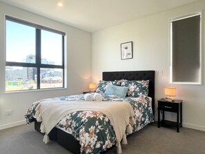 A bedroom with a large window, a bed with floral bedding, a nightstand with a lamp, and a framed picture on the wall.