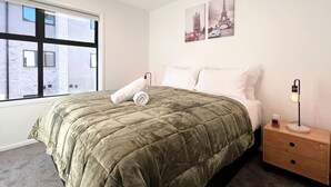 Une chambre à coucher avec un grand lit, une table de chevet, une lampe et une fenêtre donnant sur des immeubles.