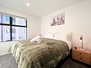 Een slaapkamer met een groot bed, een nachtkastje, een lamp en een raam met uitzicht op gebouwen.