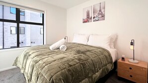 A bedroom with a large bed, a nightstand, a lamp, and a window with a view of buildings.