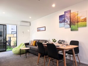 Un salón moderno con un sofá, una mesa de comedor con sillas y un sillón de espuma verde. Hay tres cuadros en la pared y una ventana con vistas al exterior.