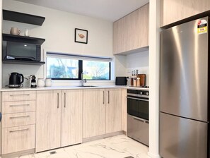 Een moderne keuken met lichte houten keukenkastjes, een roestvrijstalen koelkast, een inbouwoven en een raam met uitzicht op bomen.