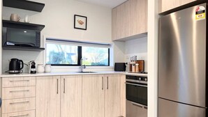 A modern kitchen with light wood cabinets, a stainless steel refrigerator, a built-in oven, and a window with a view of trees.