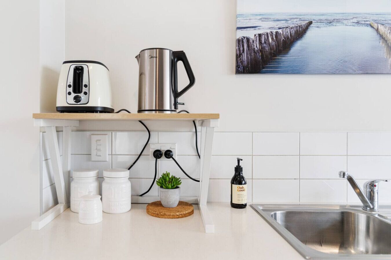 Una cocina moderna con una tetera de acero inoxidable, un tostador y una cafetera sobre una encimera de madera. Hay una planta en maceta, una botella de aceite de oliva y una taza blanca en la encimera. Se ve un fregadero a la derecha y un cuadro de un muelle en la pared.
