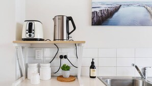 Eine moderne Küche mit einem Edelstahl-Wasserkocher, einem Toaster und einer Kaffeemaschine auf einer Holz Arbeitsplatte. Auf der Arbeitsplatte stehen ein Topfpflanze, eine Flasche Olivenöl und eine weiße Tasse. Rechts ist ein Spülbecken zu sehen, an der Wand hängt ein Gemälde von einem Steg.