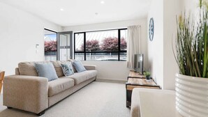 Ein modernes Wohnzimmer mit einem beigefarbenen Sofa, einem Holztisch und einem großen Fenster mit Blick auf Bäume.