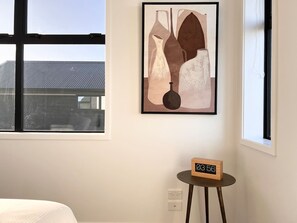 A room with a bed, a wall-mounted abstract painting, a small table with a clock, and a window with a view of a building. - Lichfield city stay with a super king suite in Christchurch (Christchurch)