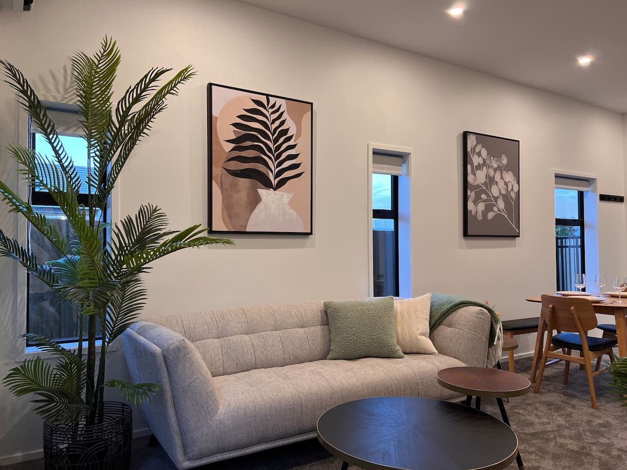 A living room with a sofa, a coffee table, and a dining table with chairs. There are two windows and a plant.