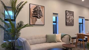 A living room with a sofa, a coffee table, and a dining table with chairs. There are two windows and a plant.