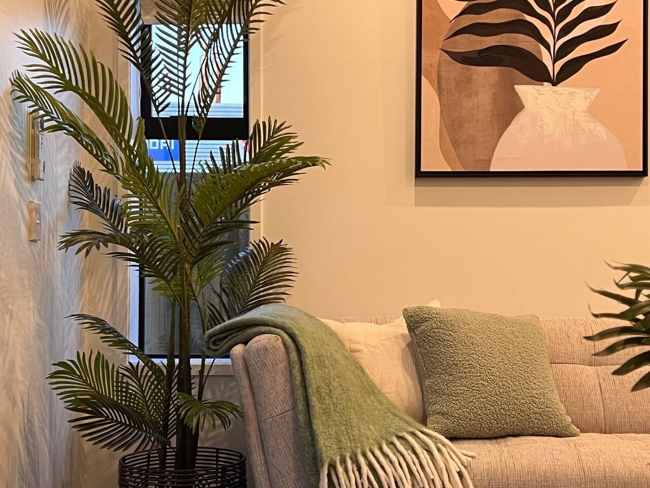 A living room with a large plant, a couch with a green throw blanket, and a framed artwork on the wall.