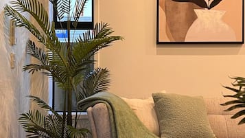 A living room with a large plant, a couch with a green throw blanket, and a framed artwork on the wall.