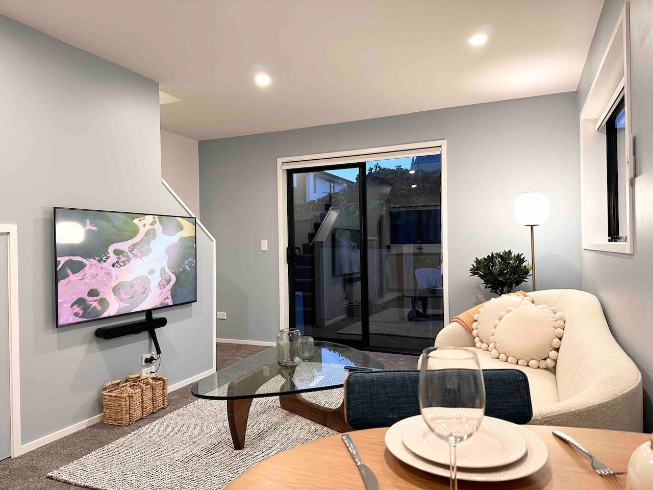 A modern living room with a flat-screen TV, a glass coffee table, a sofa with decorative pillows, and a dining area with a table set for two.