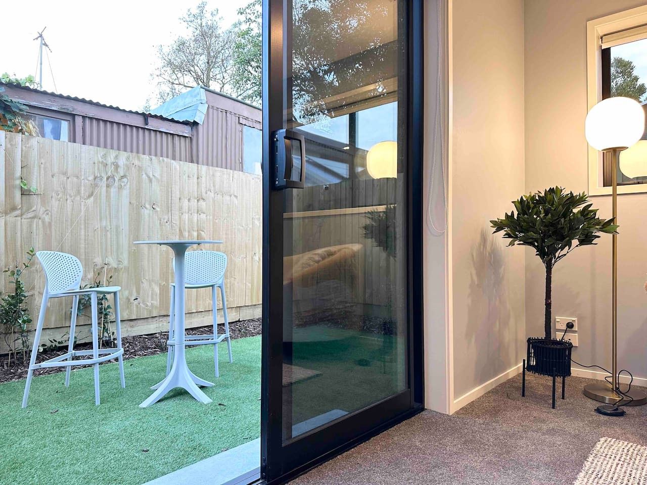 A room with a sliding glass door leading to a backyard with artificial grass, two white bar stools, and a potted plant.