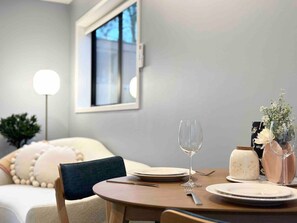A dining area with a round table set for two, a vase with flowers, and a window with a view.