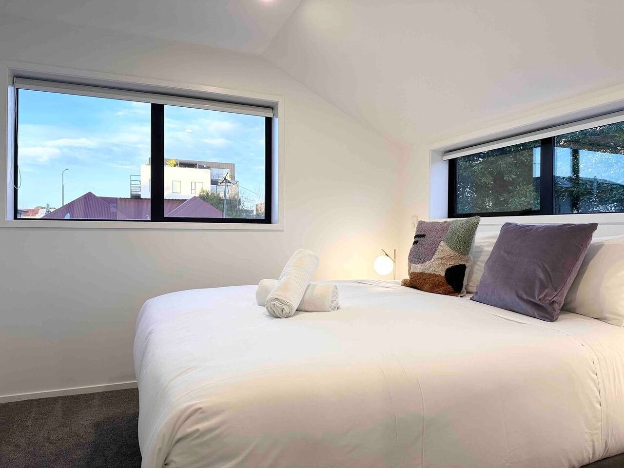 Ein modernes Schlafzimmer mit einem großen Bett, zwei Fenstern und Blick auf Gebäude.