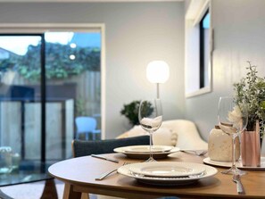 Una mesa de comedor servida con platos, cubiertos y copas de vino, con un florero y una lámpara al fondo.
