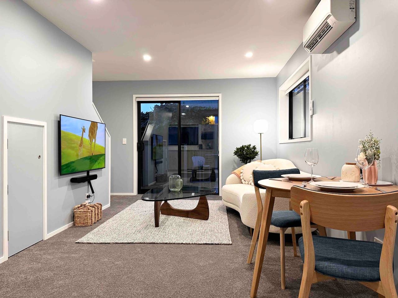 A modern living room with a flat-screen TV mounted on the wall, a glass coffee table, a sofa, and a dining table set for two.